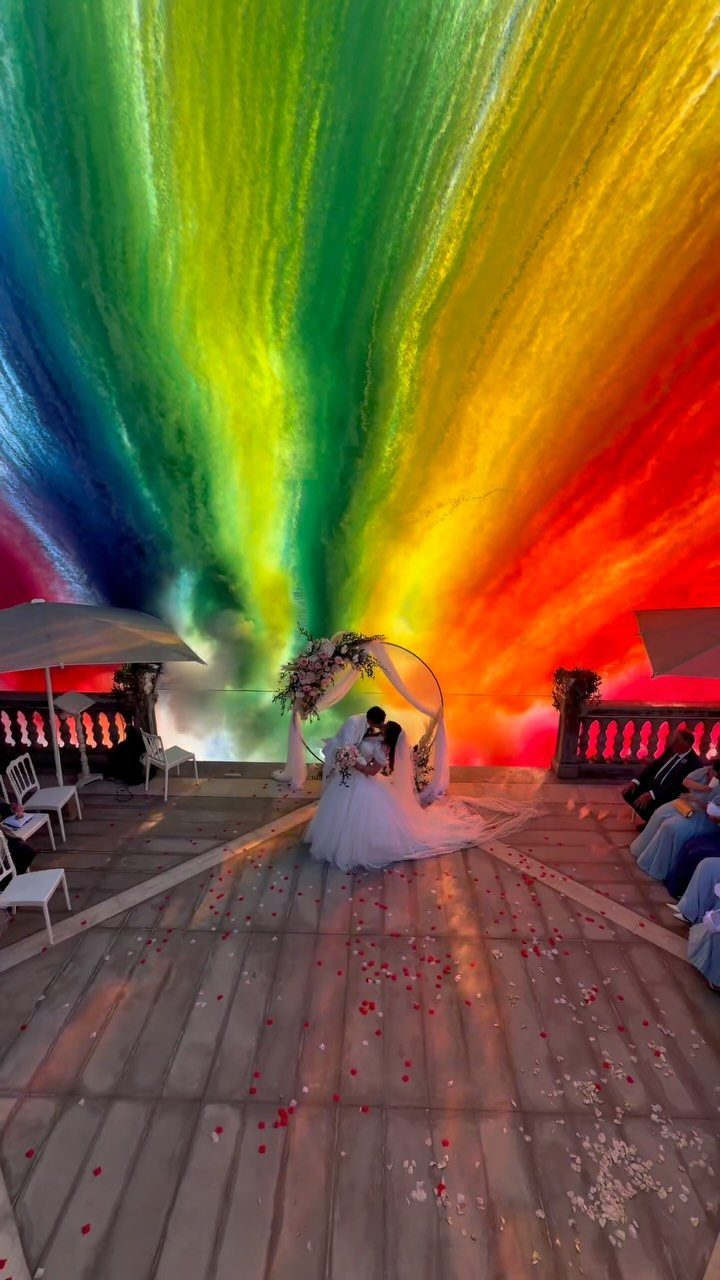 Bride and groom kissing under a floral arch with a giant rainbow smoke explosion.