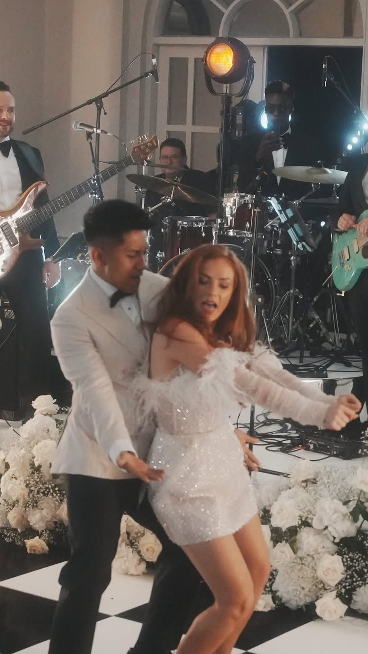 Couple dancing in formal white attire with a live band and white flowers.