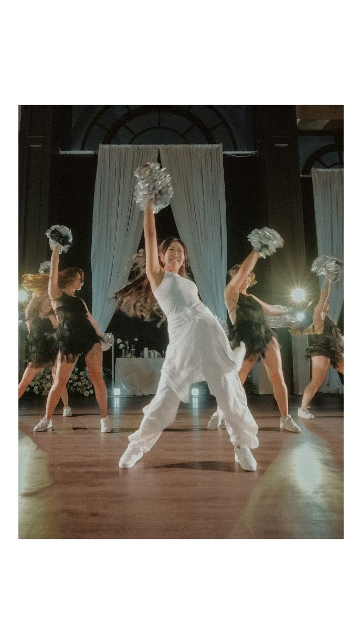 Bride in white jumpsuit dancing with bridesmaids using silver pom-poms at a wedding.