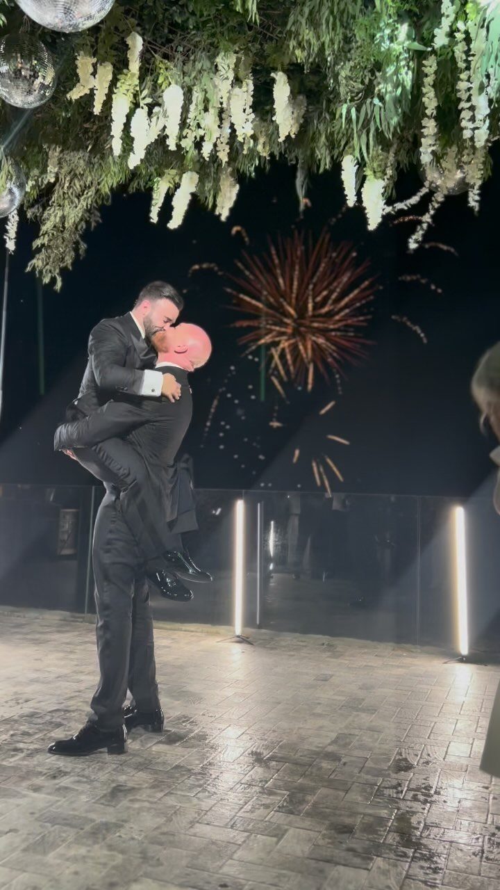 Two men in suits kissing on a dance floor with fireworks in the background.