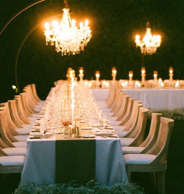 Long outdoor dinner table with white linens, glowing candles, and crystal chandeliers.