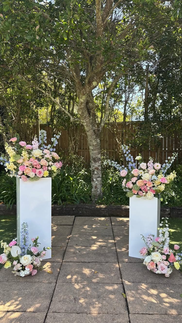 Floral arrangements in soft blue, pink, and white on white pedestals outdoors.