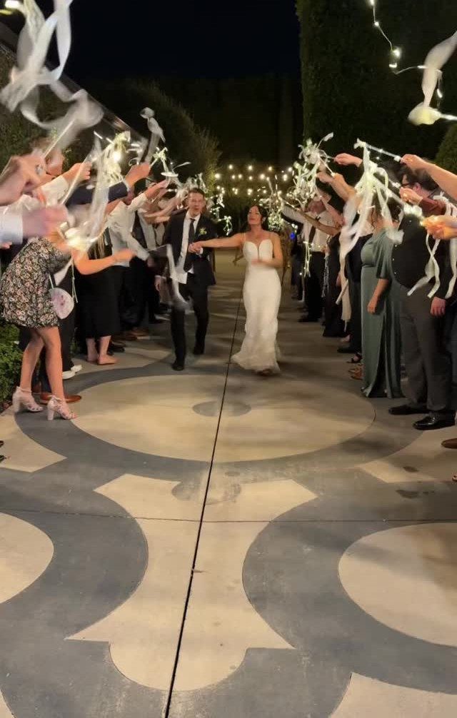 Bride and groom walking through a crowd of guests waving white ribbons at night.