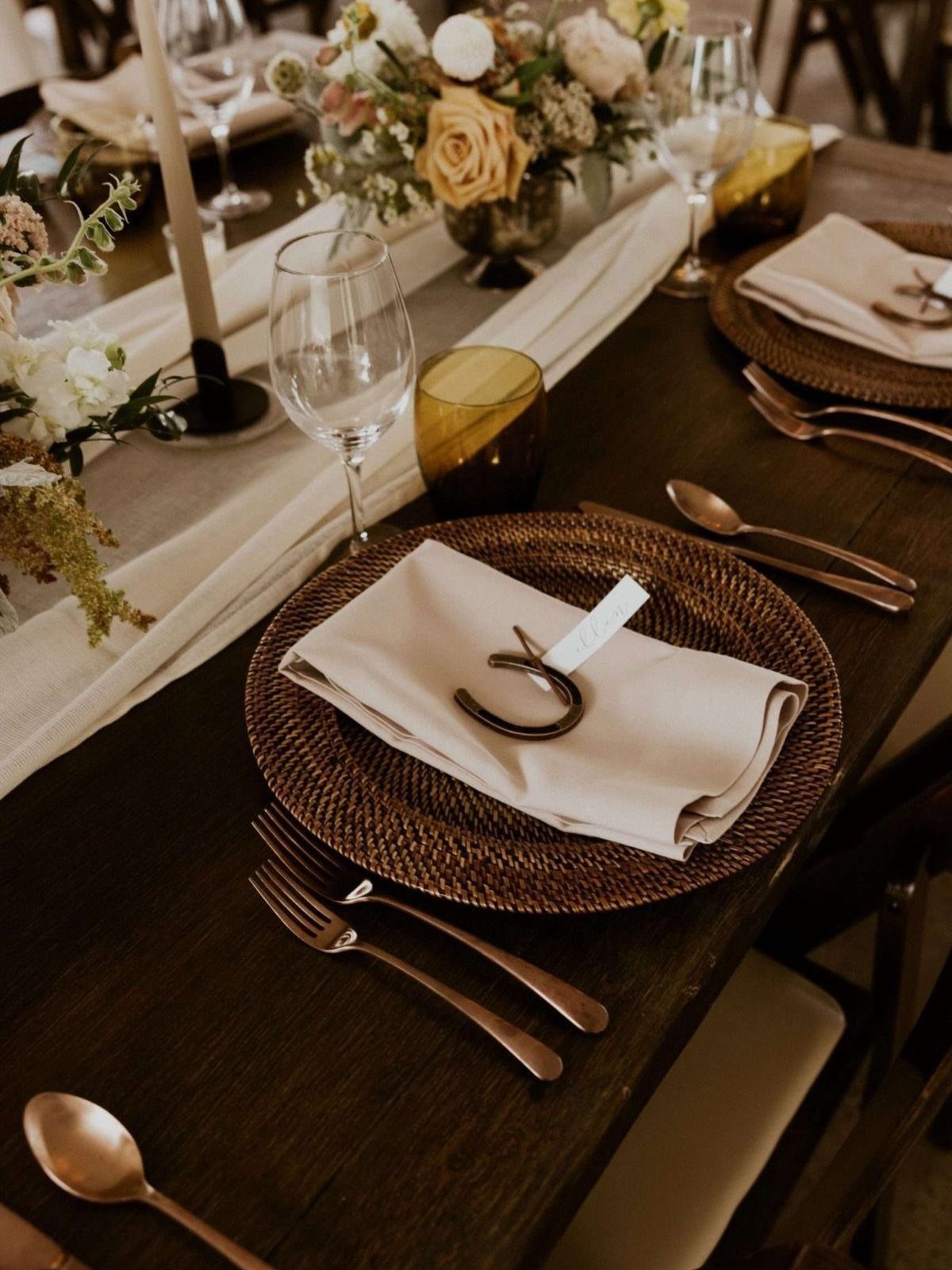 Rustic wedding table setting with woven chargers, copper cutlery, and horseshoe napkin rings.