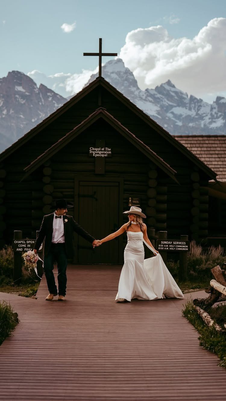 Bride and groom in cowboy hats holding hands before a rustic log chapel.