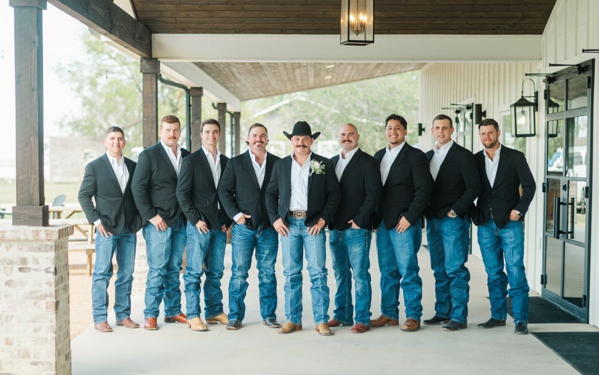 Nine men in black blazers and blue jeans posing under a porch.