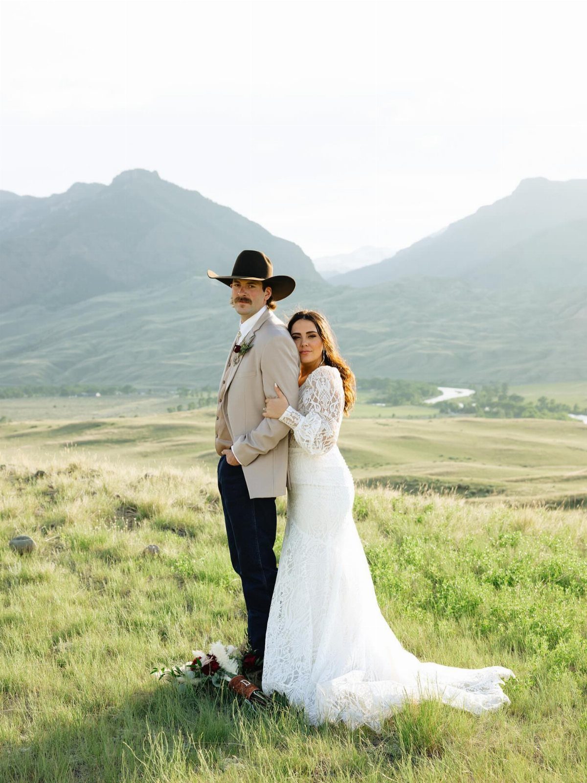 Groom in western attire and bride in lace dress posing in mountain meadow.