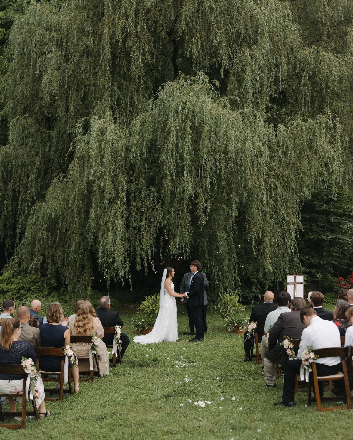 Bride and groom exchanging vows under a large weeping willow tree outdoors.