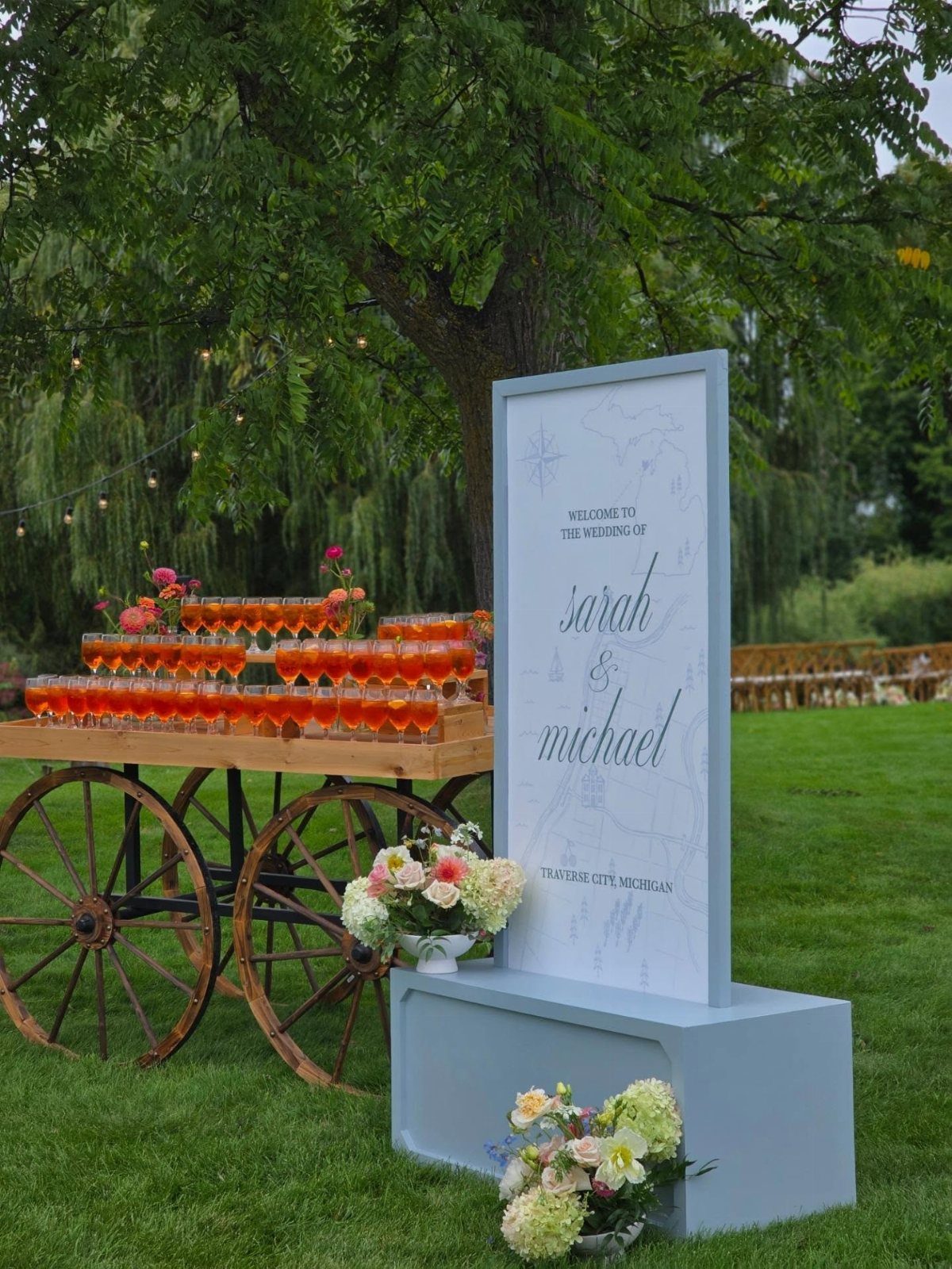 Wedding welcome sign and drink cart on a lawn under a willow tree