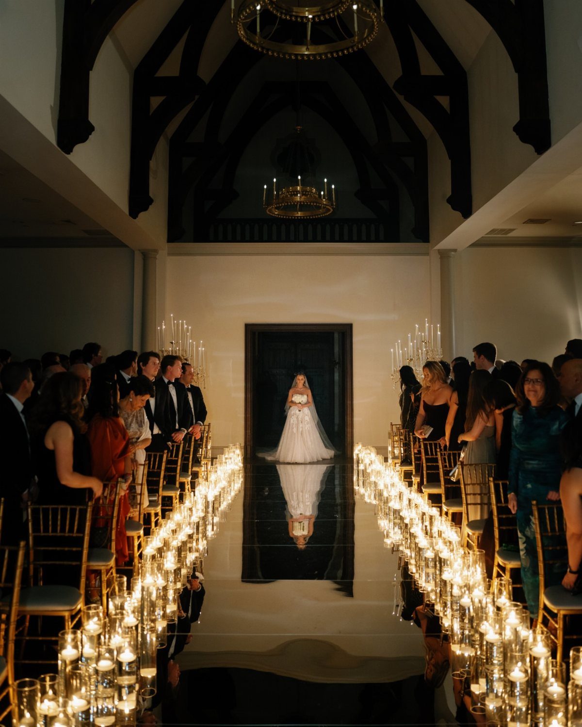 Bride walking down a black aisle lined with glowing candles in glass cylinders.