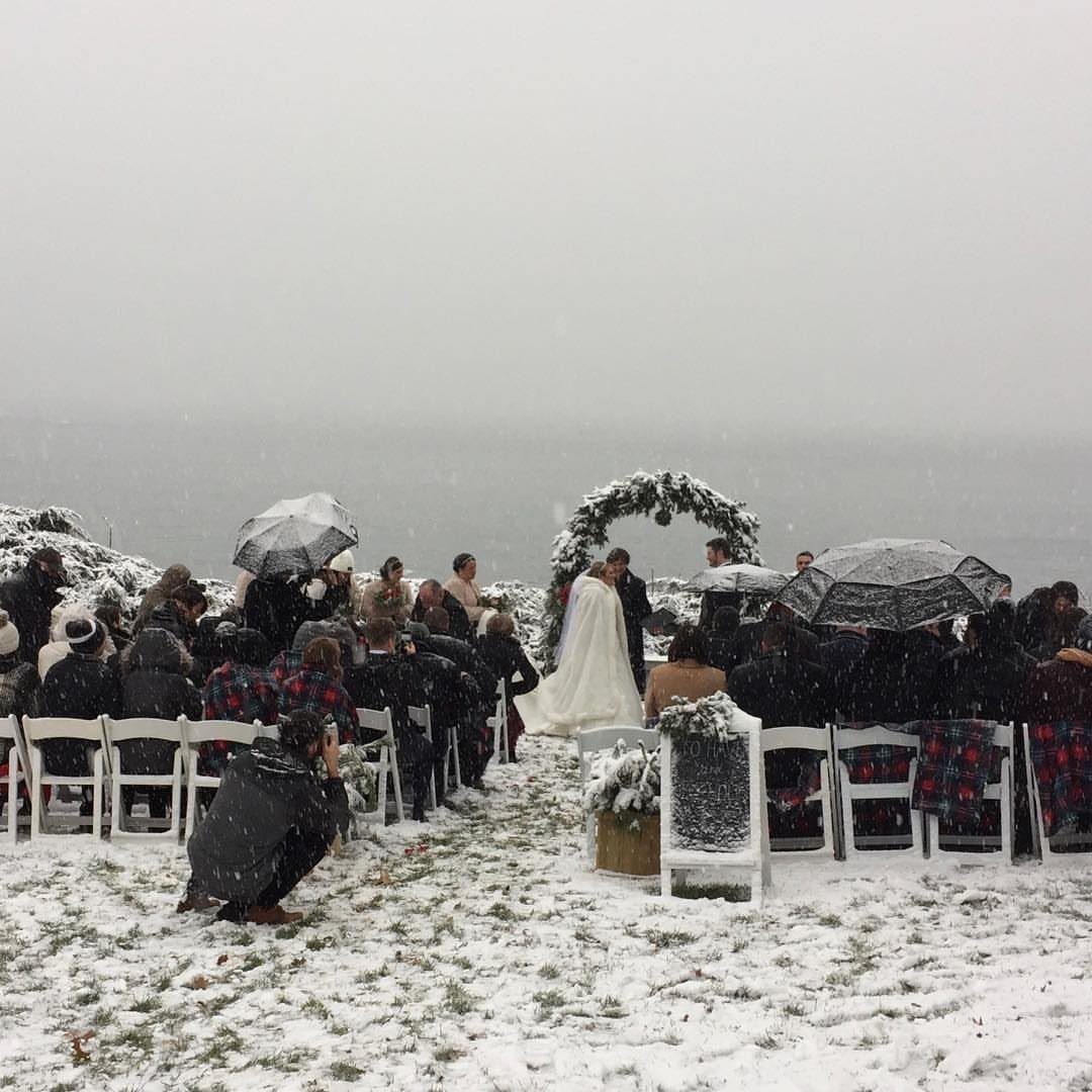 Wedding ceremony in the snow with a greenery arch and guests with umbrellas.
